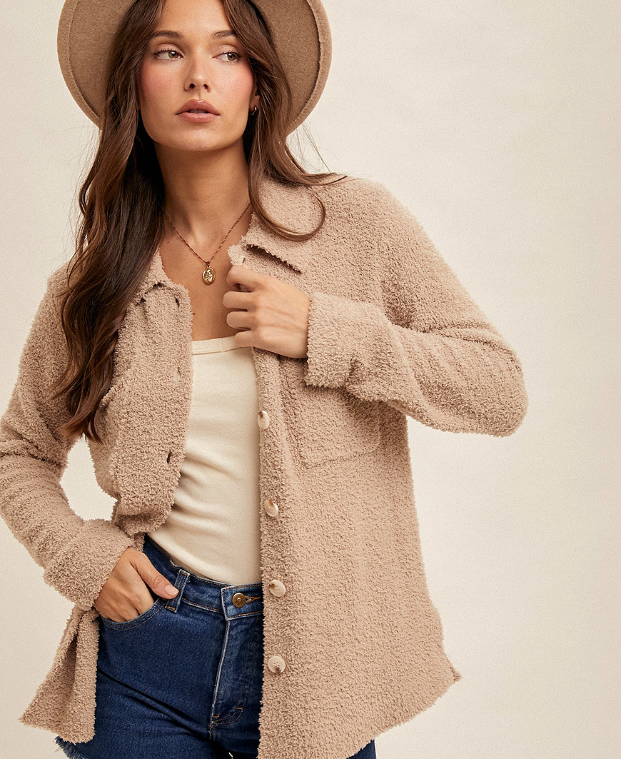 Woman wearing a brown cardigan, white top, and blue jeans with a beige hat on a plain background