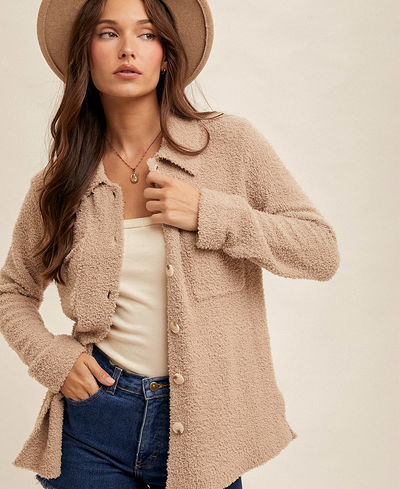 Woman wearing a brown cardigan, white top, and blue jeans with a beige hat on a plain background