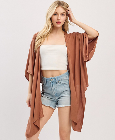 Woman wearing a brown kimono over a white top and denim shorts on a light gray background