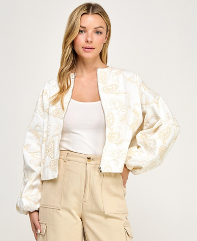 Woman wearing a white patterned jacket over a white top and beige pants on a light gray background