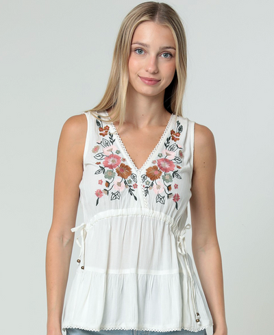 Woman wearing a white sleeveless top with floral embroidery on a plain background